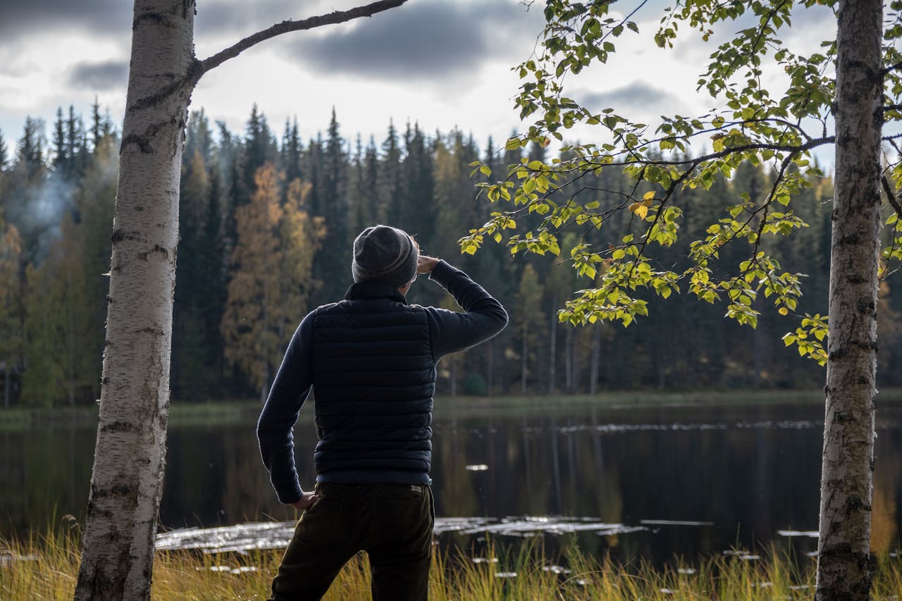A man stands by a serene lake in Finland, surrounded by autumn foliage, enjoying the peaceful forest scenery.